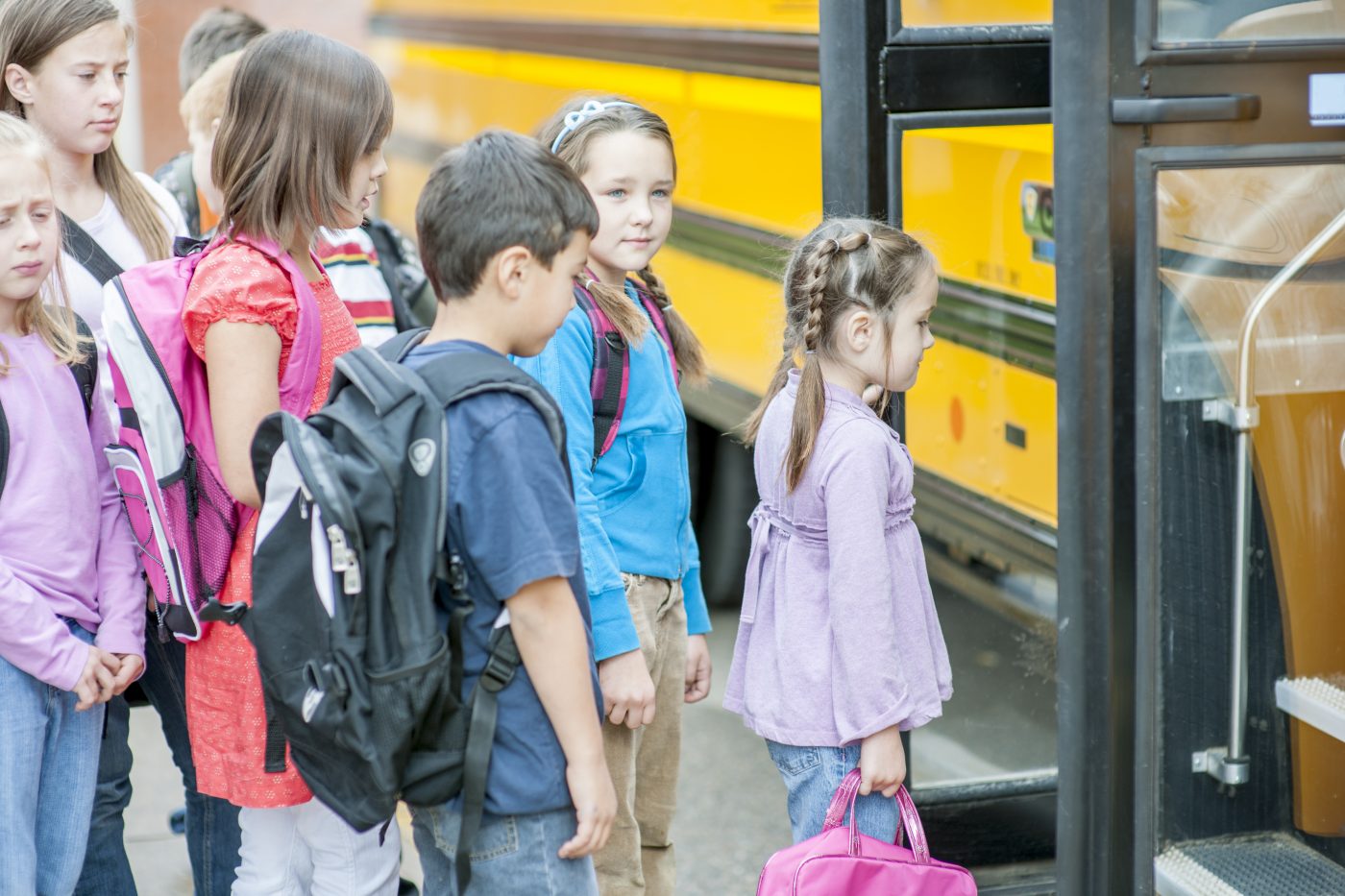 A multi-ethnic group of elementary age children are standing outside of a school bus and are waiting to get on.